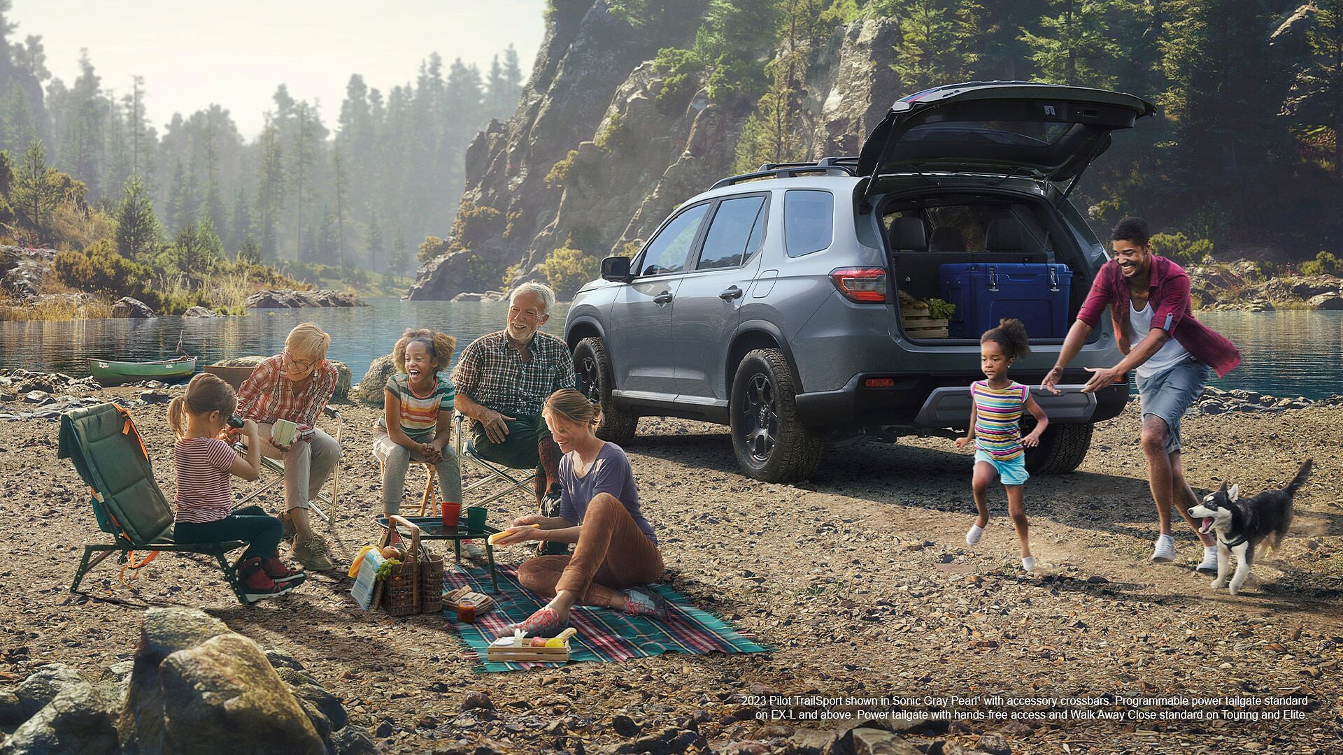 A family having a picnic on the lake shore behind them is a 2023 Honda Pilot TrailSport shown in Sonic Gray Pearl with accessory crossbars. Programmable power tagilgate standard on EX-L and aboce. Power tailgate with hands-free aaccess and Walk Away Close standard on Touring and Elite